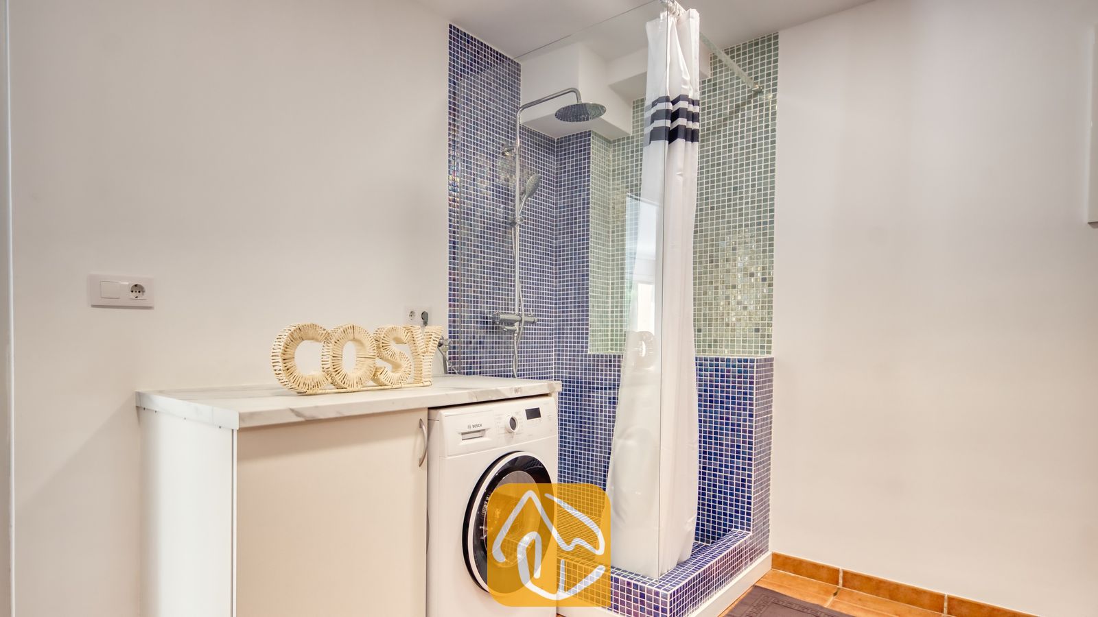 Vakantievilla's Costa Brava Spanje - Villa La Luna - Badkamer met een witte wasmachine naast een aanrechtblad met een geweven 'cosy'-bordje, en een doucheruimte voorzien van blauwe mozaïektegels.