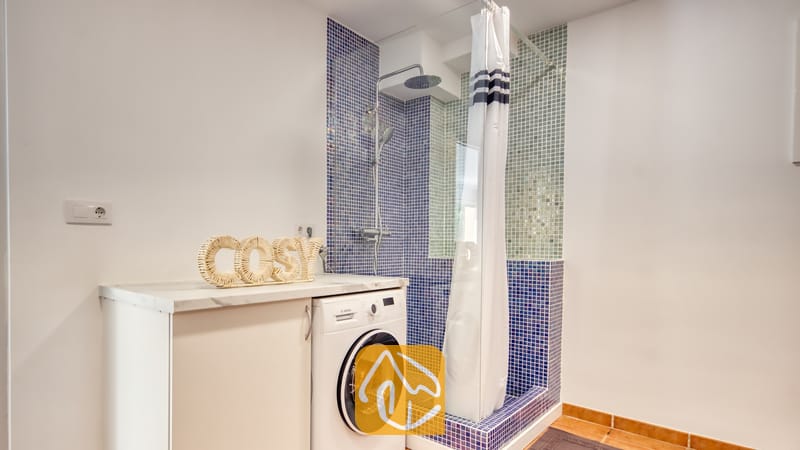Holiday villas Costa Brava Spain - Villa La Luna - Bathroom featuring a white washing machine next to a counter with a woven cosy sign, and a shower area with blue mosaic tiles.