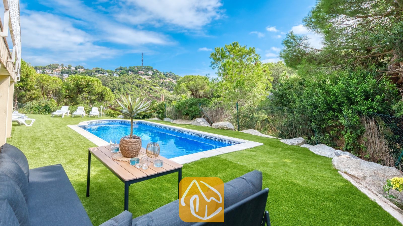 Vakantievilla's Costa Brava Spanje - Villa La Luna - Privézwembad en tuin bij Villa La Luna, voorzien van een rechthoekig zwembad, een houten salontafel met een in een pot geplante plant, een grijze buitenbank en omringende groene bomen onder een blauwe hemel.
