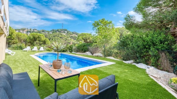 Vakantievilla's Costa Brava Spanje - Villa La Luna - Privézwembad en tuin bij Villa La Luna, voorzien van een rechthoekig zwembad, een houten salontafel met een in een pot geplante plant, een grijze buitenbank en omringende groene bomen onder een blauwe hemel.