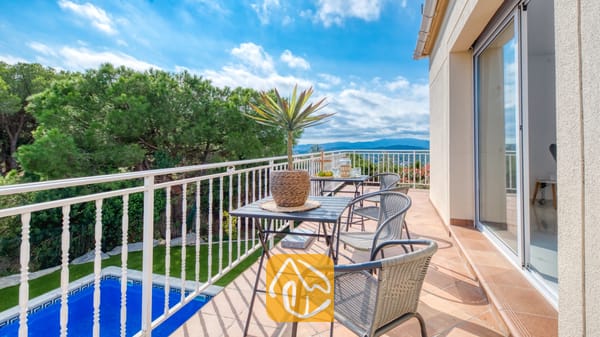 Vakantievilla's Costa Brava Spanje - Villa La Luna - Terras van Villa La Luna met een buitenetablissement, een potplant en een witte reling met uitzicht op een zwembad en bomen.