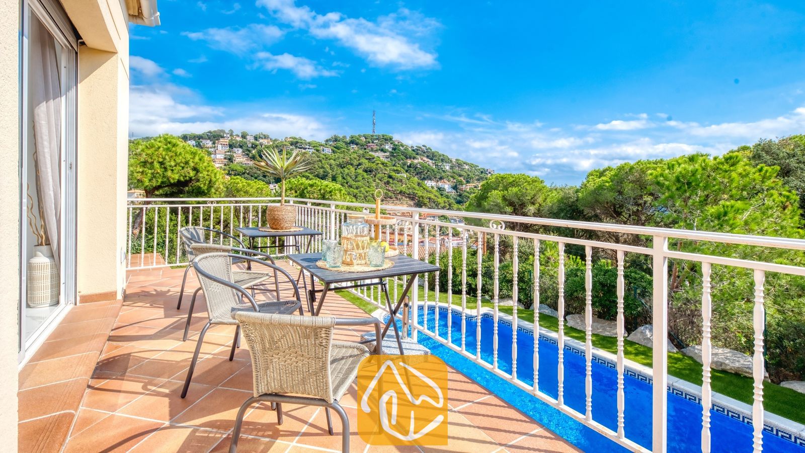 Vakantievilla's Costa Brava Spanje - Villa La Luna - Zonnig balkon bij Villa La Luna met betegelde vloer, een eettafel met gevlochten stoelen en een wit metalen hekwerk dat uitkijkt over een zwembad en groene heuvels.