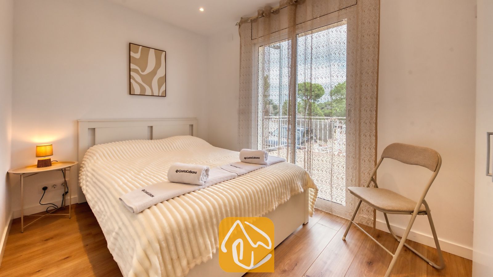 Vakantievilla's Costa Brava Spanje - Villa La Luna - Slaapkamer met een tweepersoonsbed voorzien van opgevouwen witte handdoeken, houten vloeren, een klein nachtkastje met een sfeervolle lamp en een glazen schuifpui met inbetween gordijnen.