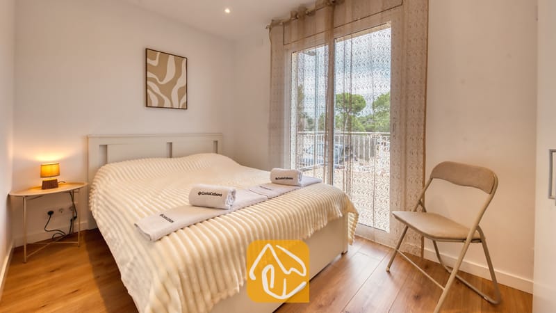 Holiday villas Costa Brava Spain - Villa La Luna - Bedroom featuring a double bed with folded white towels, wooden flooring, a small bedside table with a glowing lamp, and a sliding glass door with sheer curtains.