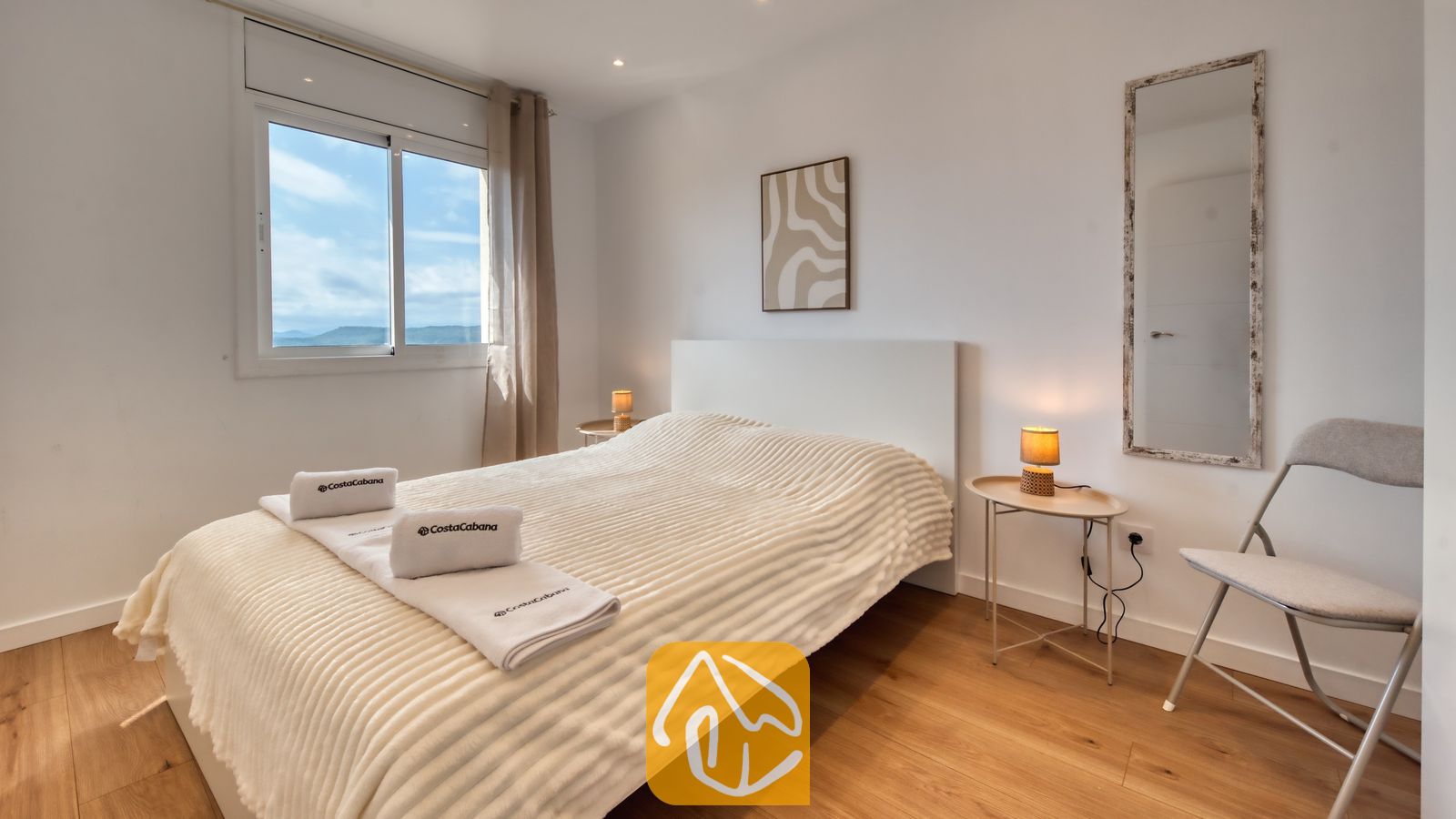 Vakantievilla's Costa Brava Spanje - Villa La Luna - Slaapkamer met een wit bed, houten vloer, nachtkastjes met kleine lampen en een raam met uitzicht op een bewolkte lucht.