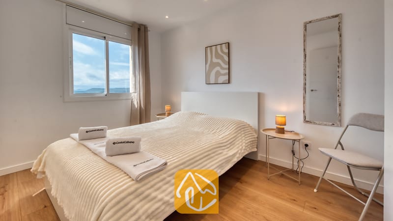 Holiday villas Costa Brava Spain - Villa La Luna - Bedroom with a white bed, wooden flooring, side tables with small lamps, and a window showing a cloudy sky.