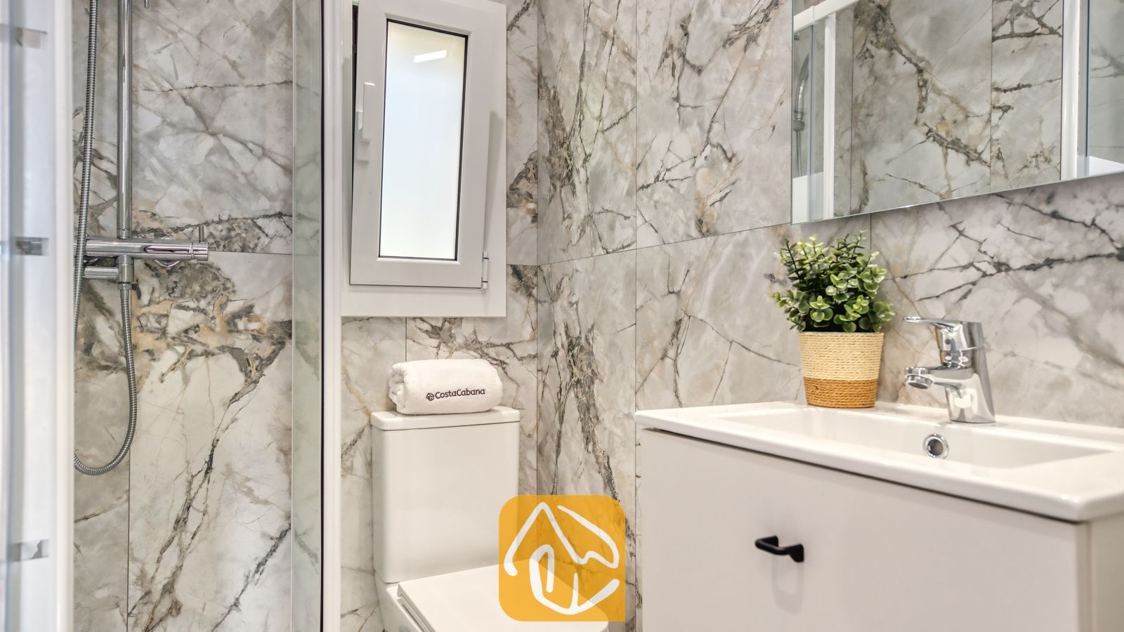 Vakantievilla's Costa Brava Spanje - Villa La Luna - Moderne badkamer met wandtegels met grijs marmer-effect, een wit toilet met een opgerolde handdoek, een witte wastafel met chromen kranen en een glazen inloopdouche.