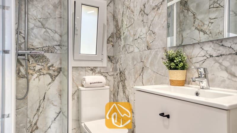 Holiday villas Costa Brava Spain - Villa La Luna - Modern bathroom featuring grey marble-effect tiled walls, a white toilet with a rolled towel, a white vanity sink with chrome fixtures, and a glass-enclosed shower.