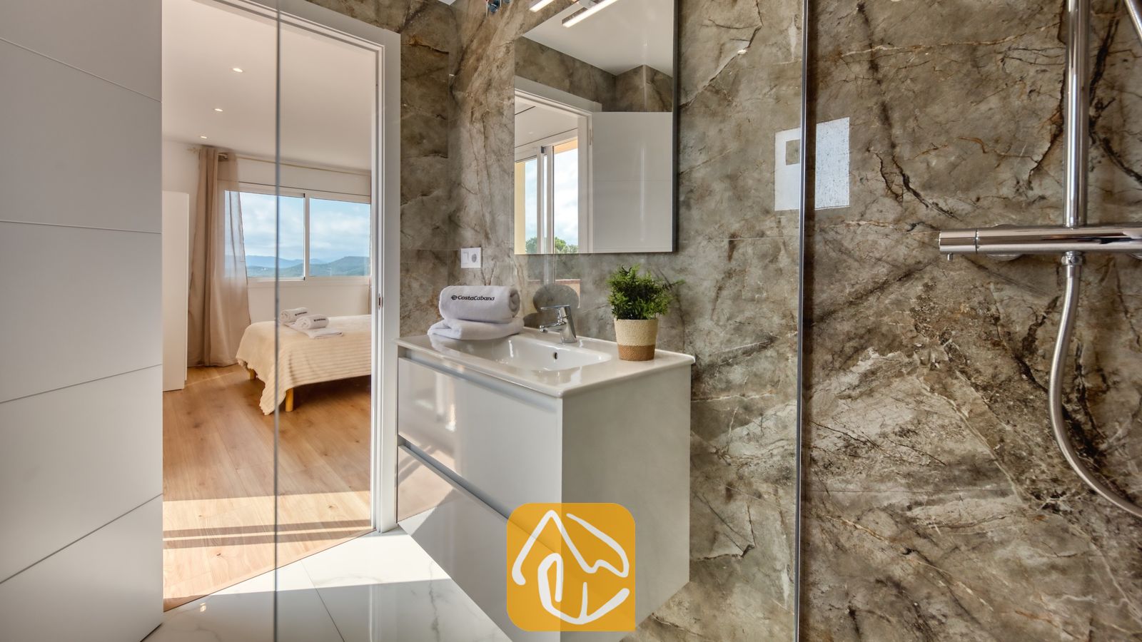 Vakantievilla's Costa Brava Spanje - Villa La Luna - Moderne badkamer met wanden in bruine marmerlook tegels, een witte wastafel met meubel, een wandspiegel en een aangrenzende slaapkamer die zichtbaar is door een open deuropening.