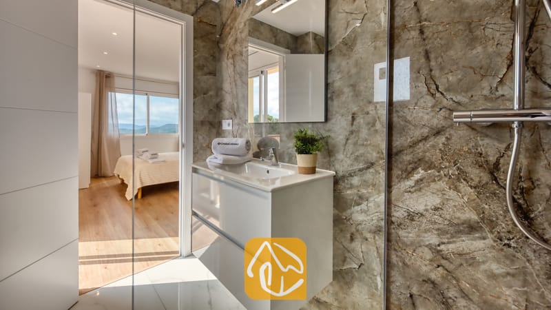Holiday villas Costa Brava Spain - Villa La Luna - Modern bathroom with brown marble-effect tile walls, a white vanity sink, a wall mirror, and an adjoining bedroom visible through an open doorway.