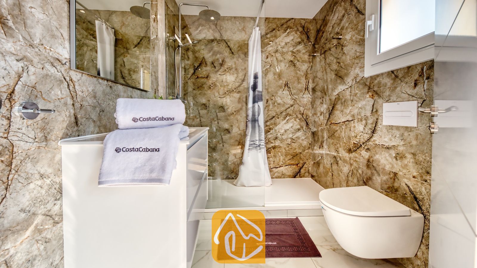 Vakantievilla's Costa Brava Spanje - Villa La Luna - Moderne badkamer met bruine marmeren muren, een wit badmeubel met opgevouwen handdoeken met logo, een inloopdouche en een hangend toilet.