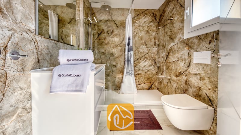 Holiday villas Costa Brava Spain - Villa La Luna - Modern bathroom featuring brown marble walls, a white vanity with folded branded towels, a walk-in shower enclosure, and a wall-mounted toilet.