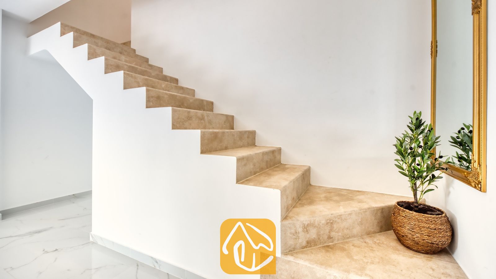 Vakantievilla's Costa Brava Spanje - Villa La Luna - Binnenstrap met treden van beige natuursteen en witte muren, verrijkt met een kamerplant in een geweven mand en een goud ingelijste spiegel onderaan de trap.
