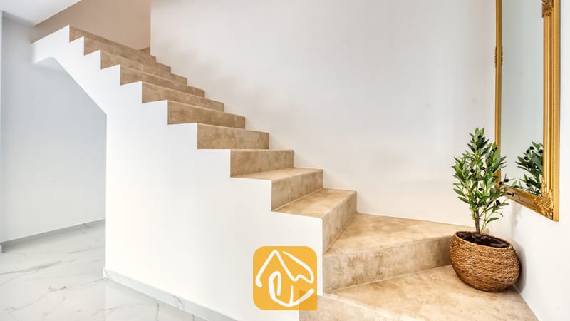 Holiday villas Costa Brava Spain - Villa La Luna - Indoor staircase with beige stone treads and white walls, featuring a potted plant in a woven basket and a gold-framed mirror at the base.