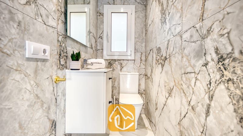 Holiday villas Costa Brava Spain - Villa La Luna - Modern bathroom interior featuring grey marble-patterned tiled walls, a white vanity unit with sink, a toilet, and a small window above.