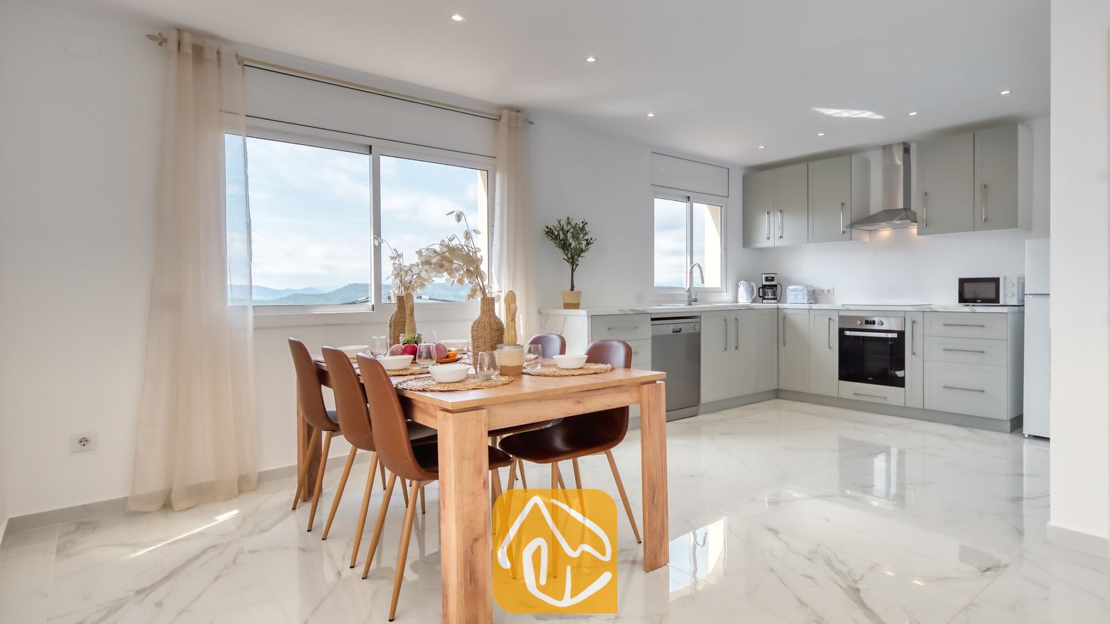Vakantievilla's Costa Brava Spanje - Villa La Luna - Houten eettafel met bruine lederen stoelen in een open keuken bij Villa La Luna, voorzien van witte marmeren tegelvloeren en grijze kasten.