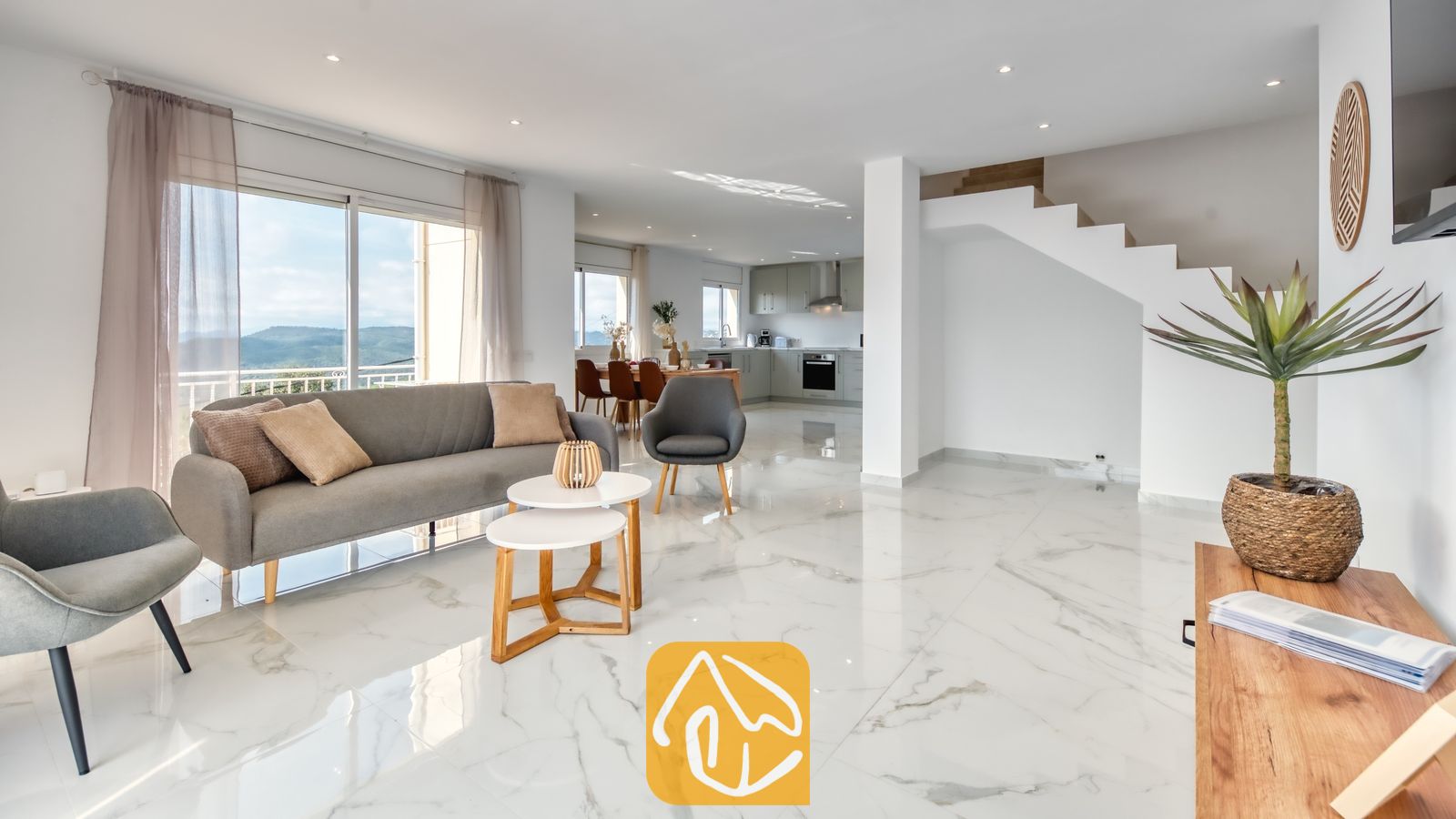 Vakantievilla's Costa Brava Spanje - Villa La Luna - De open woonkamer van Villa La Luna is voorzien van wit marmeren tegelvloeren, een grijze bank, twee bijpassende fauteuils en glazen schuifdeuren die opengaan naar een uitzicht op de heuvels.
