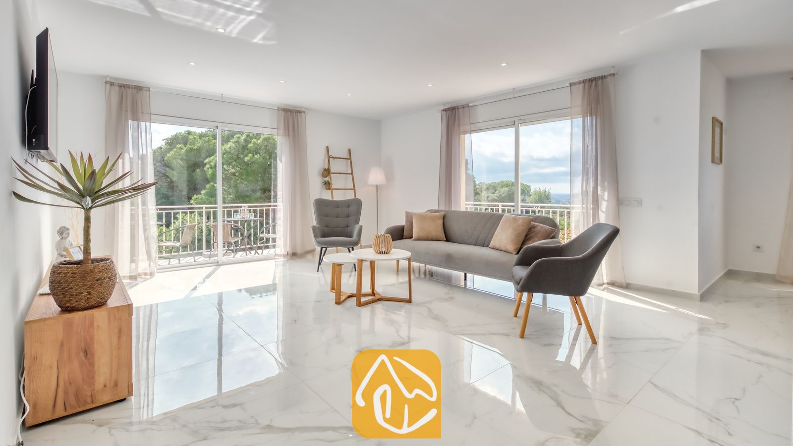 Vakantievilla's Costa Brava Spanje - Villa La Luna - Lichte woonkamer in Villa La Luna met wit marmeren vloeren, een grijze bank, bijpassende fauteuils en grote glazen schuifdeuren die opengaan naar een balkon met uitzicht op de bomen.