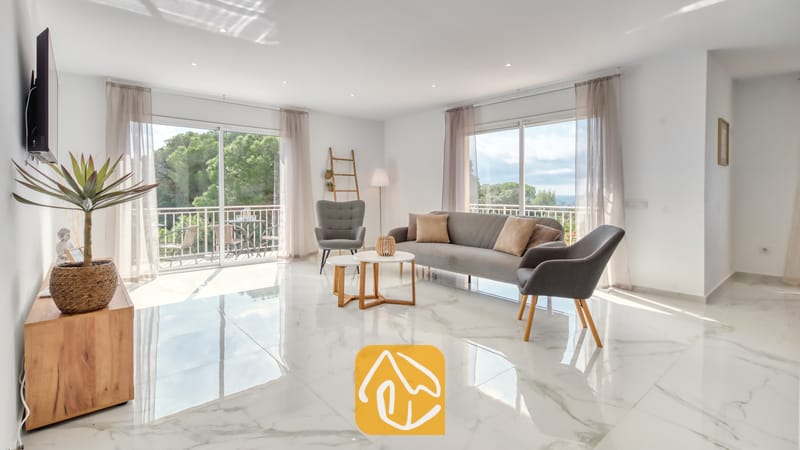 Vakantievilla's Costa Brava Spanje - Villa La Luna - Lichte woonkamer in Villa La Luna met wit marmeren vloeren, een grijze bank, bijpassende fauteuils en grote glazen schuifdeuren die opengaan naar een balkon met uitzicht op de bomen.
