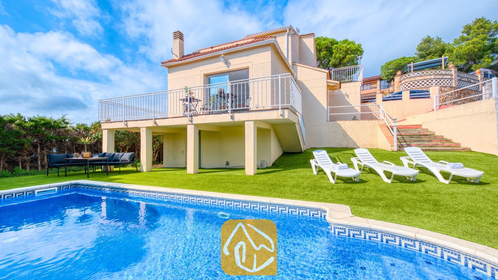 Vakantievilla's Costa Brava Spanje - Villa La Luna - Buitenkant van Villa La Luna met een privézwembad, witte ligstoelen op kunstgras en een beige huis van twee verdiepingen met een balkon onder een licht bewolkte hemel.