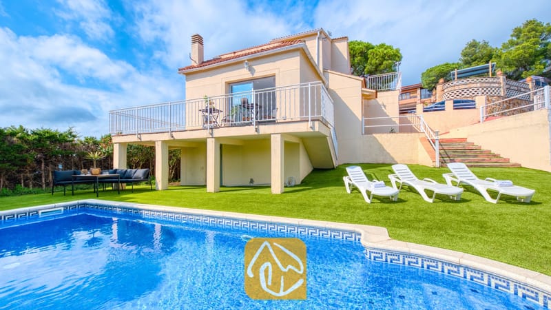 Vakantievilla's Costa Brava Spanje - Villa La Luna - Buitenkant van Villa La Luna met een privézwembad, witte ligstoelen op kunstgras en een beige huis van twee verdiepingen met een balkon onder een licht bewolkte hemel.