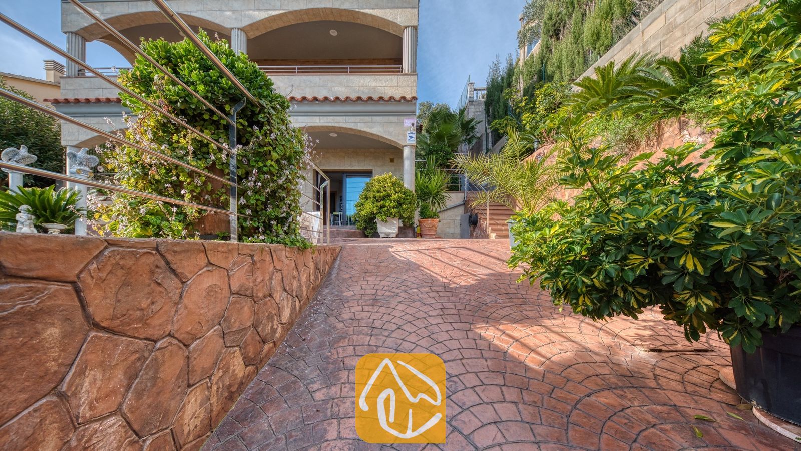 Vakantievilla's Costa Brava Spanje - Villa Grace - Een verharde terracotta oprijlaan leidt naar de multiverdiepingen tellende, beige stenen buitenkant van Villa Grace, geflankeerd door stenen muren en groene planten in potten.