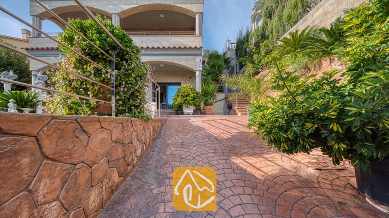 Vakantievilla's Costa Brava Spanje - Villa Grace - Een verharde terracotta oprijlaan leidt naar de multiverdiepingen tellende, beige stenen buitenkant van Villa Grace, geflankeerd door stenen muren en groene planten in potten.