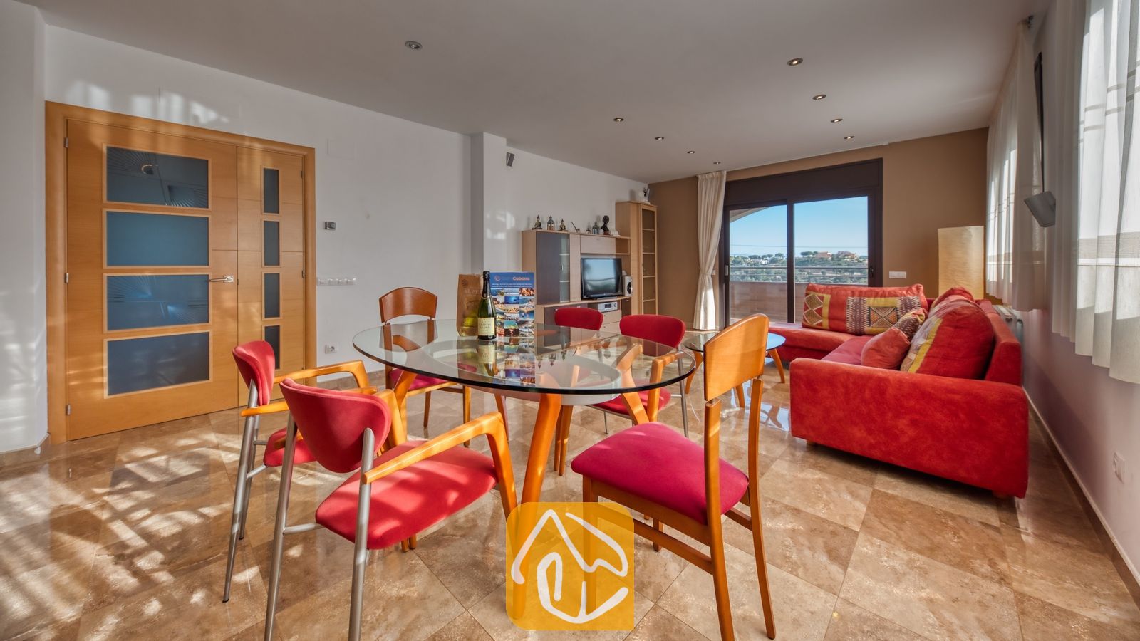 Vakantievilla's Costa Brava Spanje - Villa Grace - Ruime woon- en eetkamer in Villa Grace, voorzien van een glazen eettafel met rode stoelen en een aangrenzende rode hoekbank.