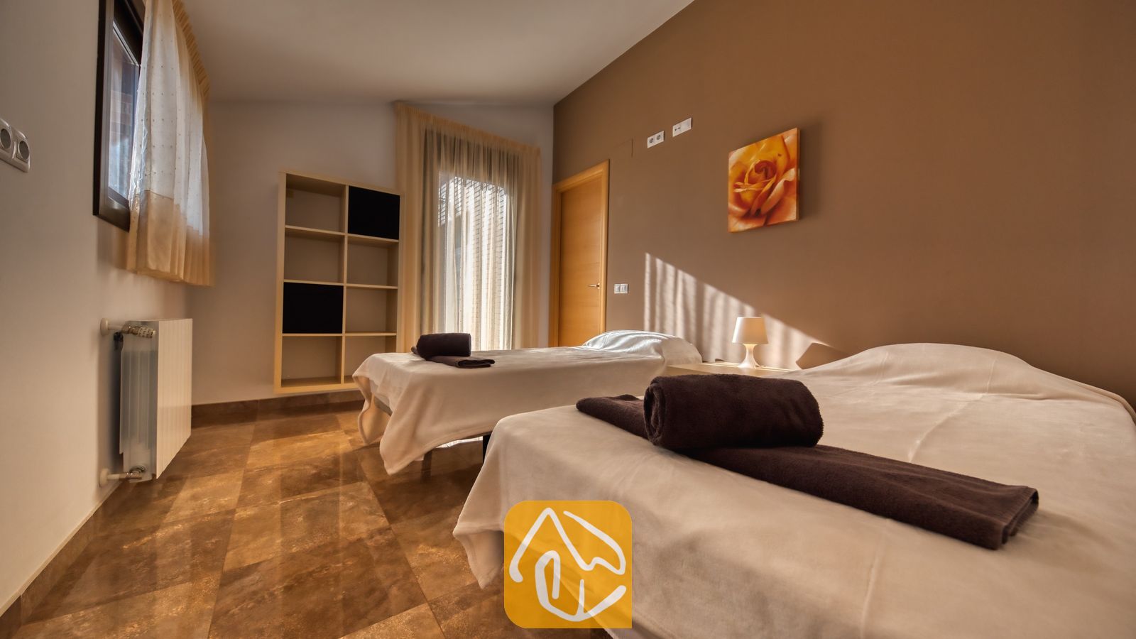 Vakantievilla's Costa Brava Spanje - Villa Grace - Tweepersoonskamer met twee eenpersoonsbedden, opgemaakt met wit beddengoed en bruine handdoeken, tegelvloeren, bruine muren en een bloemenschilderij aan de muur.