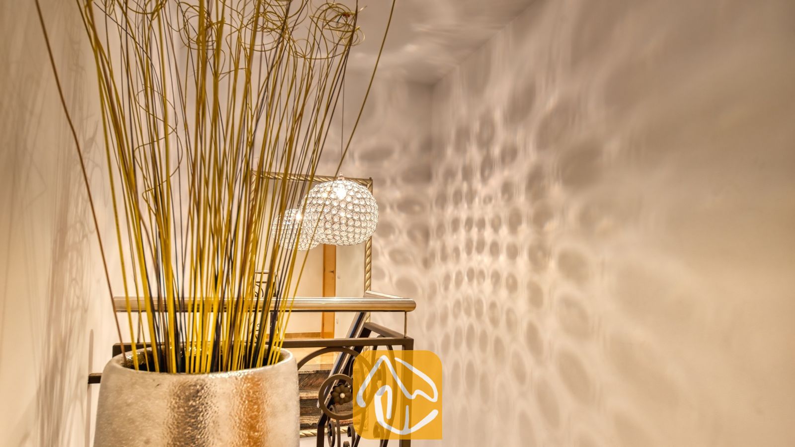 Vakantievilla's Costa Brava Spanje - Villa Grace - Decoratieve vaas met lang gedroogd gras op de voorgrond van een hal met een metalen trapleuning, een gespiegelde wand en een patroon van lichtreflecties op de witte muur.