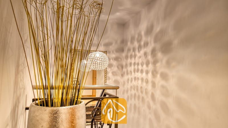 Villas de vacances Costa Brava Espagne - Villa Grace - Vase décoratif garni de hautes herbes séchées au premier plan d’un couloir doté d’une rampe d’escalier en métal, d’un mur en miroir et de reflets lumineux à motifs sur le mur blanc.