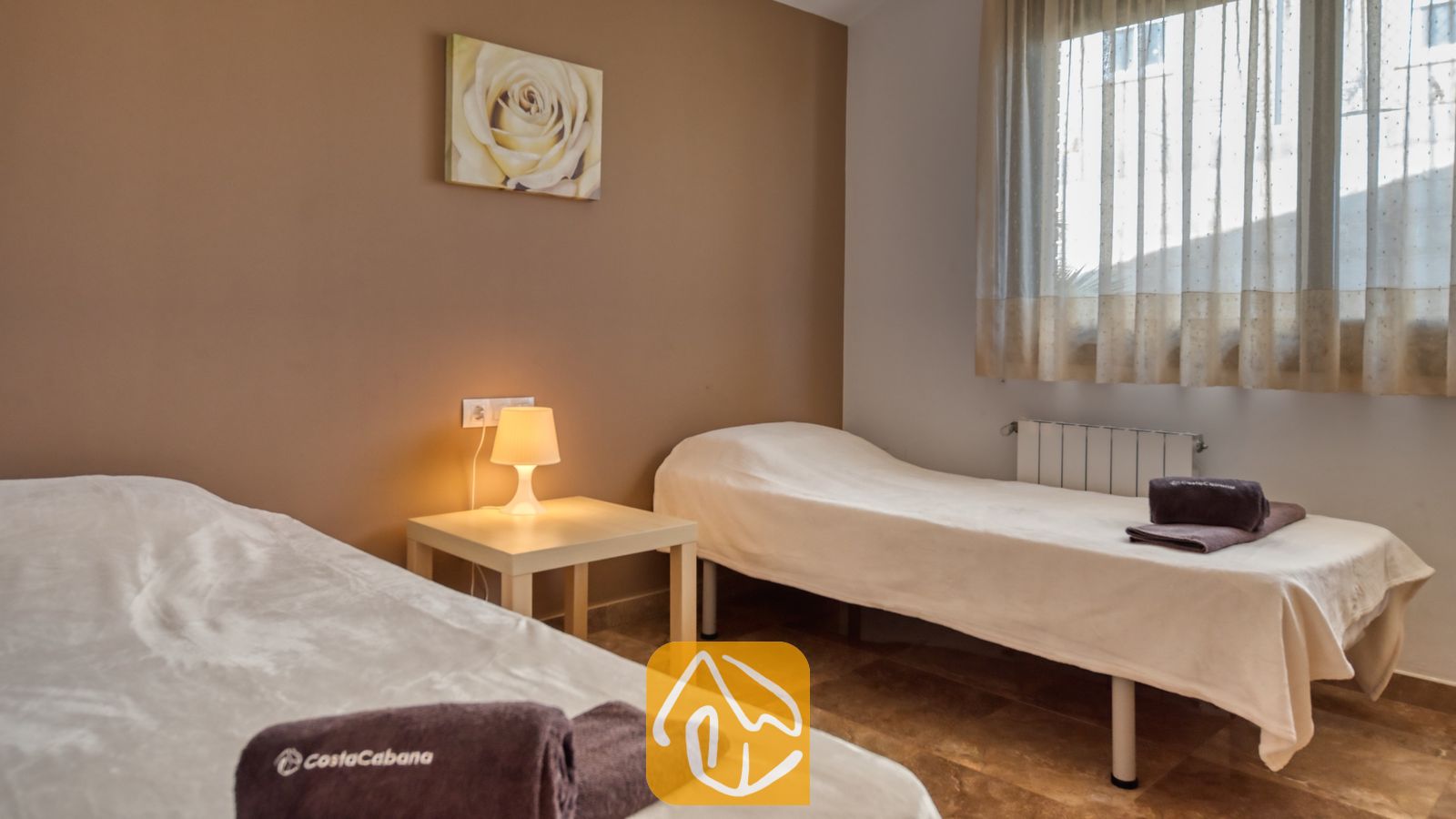 Vakantievilla's Costa Brava Spanje - Villa Grace - Tweepersoonskamer met twee eenpersoonsbedden opgemaakt met wit beddengoed, een gedeeld nachtkastje met een klein lampje en bruine wanddecoratie in Villa Grace.