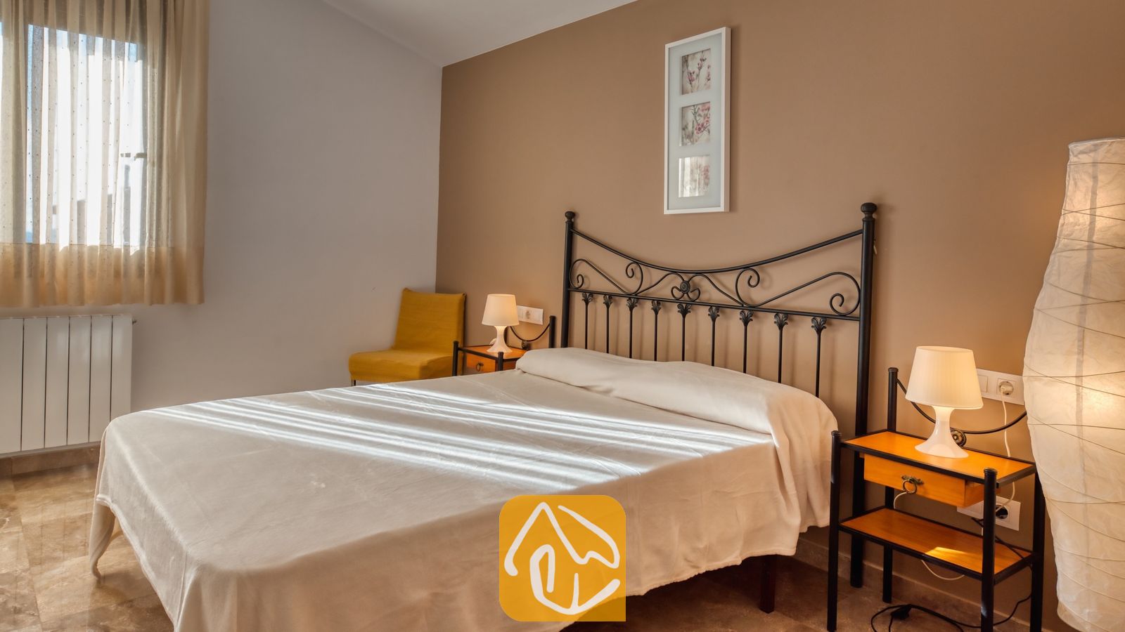 Vakantievilla's Costa Brava Spanje - Villa Grace - Slaapkamer met een zwart metalen bedframe, wit beddengoed, twee kleine nachtkastjes met lampen en een gele stoel in de hoek
