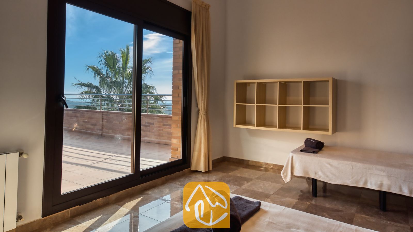 Vakantievilla's Costa Brava Spanje - Villa Grace - Slaapkamer met twee eenpersoonsbedden, voorzien van donkergerolde handdoeken, marmeren tegelvloeren, een wandboekenplank en grote glazen schuifpuien die uitkomen op een zonnig terras met uitzicht op een palmboom.