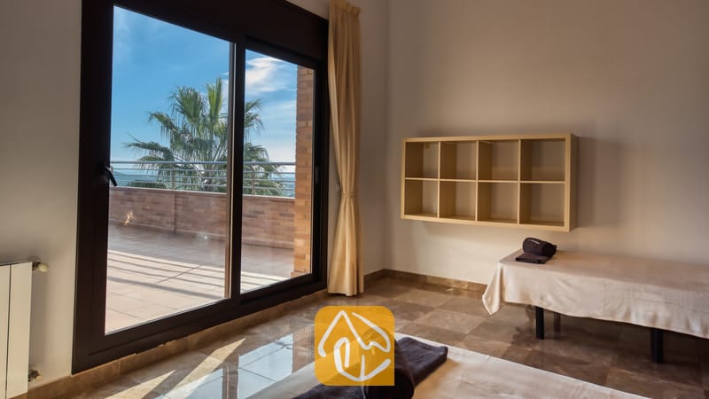 Villas de vacances Costa Brava Espagne - Villa Grace - Chambre avec deux lits simples agrémentée de serviettes sombres roulées, d'un sol en marbre, d'une étagère murale et de grandes portes coulissantes en verre s'ouvrant sur une terrasse ensoleillée avec vue sur un palmier.