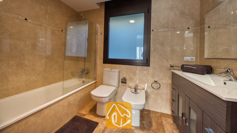 Vakantievilla's Costa Brava Spanje - Villa Grace - Badkamer met beige marmeren wandtegels, een ligbad met glazen scherm, een wit toilet, bidet en een wastafelmeubel van donker hout.