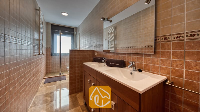 Villas de vacances Costa Brava Espagne - Villa Grace - Salle de bain équipée d'une double vasque sur meuble en bois, de deux lavabos blancs et de carrelage mural marron. Un grand miroir est suspendu au-dessus des vasques, et une douche vitrée est visible au fond.