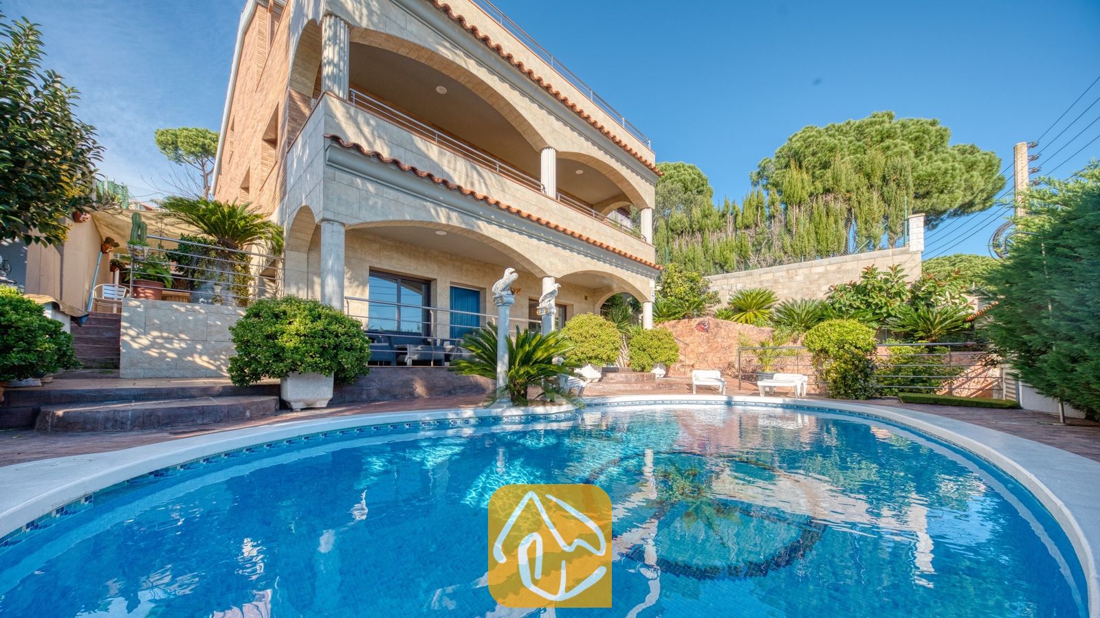 Vakantievilla's Costa Brava Spanje - Villa Grace - Gebogen zwembad met een mozaïektegelvloer, gelegen voor de meeverdiepingsbuitenkant van Villa Grace, voorzien van stenen bogen en balkons onder een helderblauwe hemel.