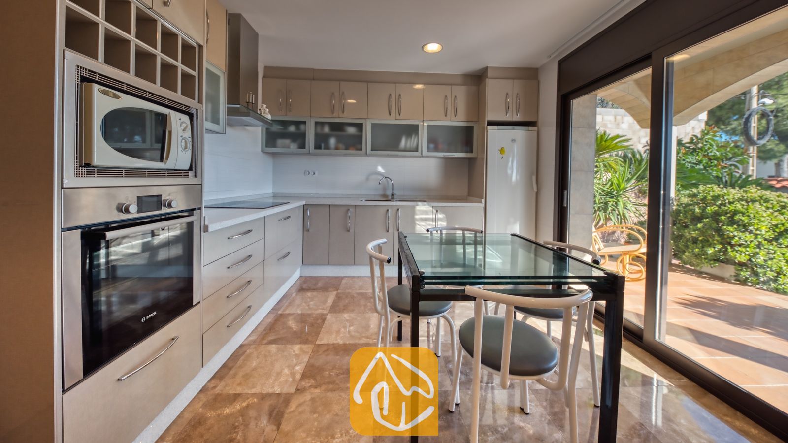 Vakantievilla's Costa Brava Spanje - Villa Grace - Keuken met beige kasten, een inbouw oven van roestvrij staal en een glazen eettafel met witte stoelen, en een schuifpui die uitkomt op een zonnig terras.