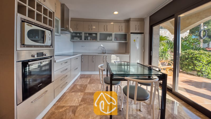 Vakantievilla's Costa Brava Spanje - Villa Grace - Keuken met beige kasten, een inbouw oven van roestvrij staal en een glazen eettafel met witte stoelen, en een schuifpui die uitkomt op een zonnig terras.