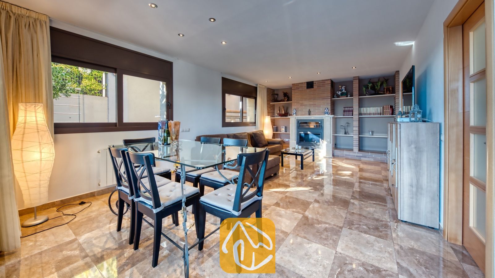Vakantievilla's Costa Brava Spanje - Villa Grace - De open woon- en eetkamer van Villa Grace is voorzien van een glazen eettafel, donkerhouten stoelen met grijze kussens, gepolijste tegelvloeren en een stenen open haard.