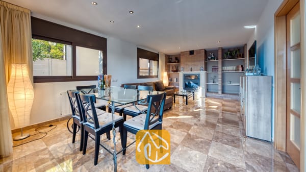 Vakantievilla's Costa Brava Spanje - Villa Grace - De open woon- en eetkamer van Villa Grace is voorzien van een glazen eettafel, donkerhouten stoelen met grijze kussens, gepolijste tegelvloeren en een stenen open haard.