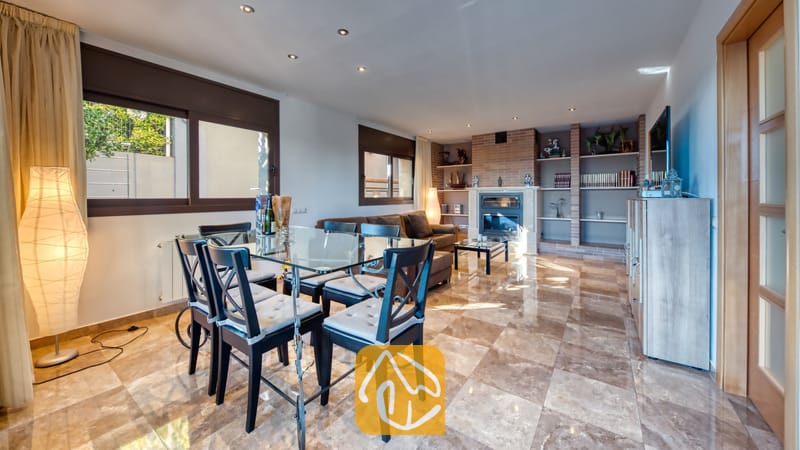 Vakantievilla's Costa Brava Spanje - Villa Grace - De open woon- en eetkamer van Villa Grace is voorzien van een glazen eettafel, donkerhouten stoelen met grijze kussens, gepolijste tegelvloeren en een stenen open haard.