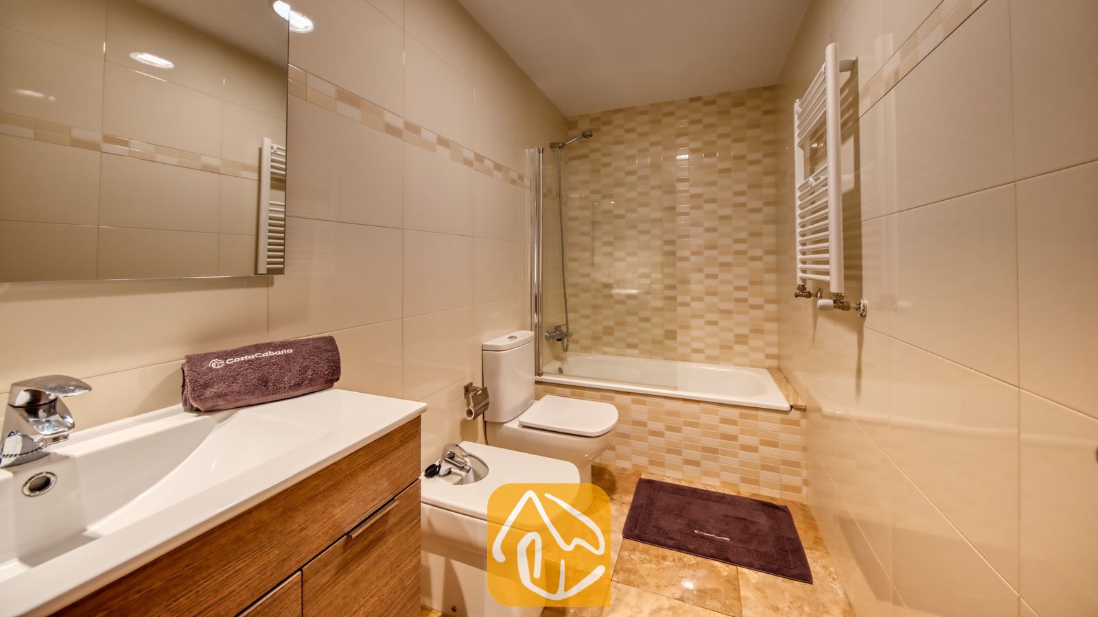 Vakantievilla's Costa Brava Spanje - Villa Grace - Moderne badkamer met beige betegelde muren, een houten badkamermeubel met een witte wastafel, toilet, bidet en een ligbad met een glazen scherm.