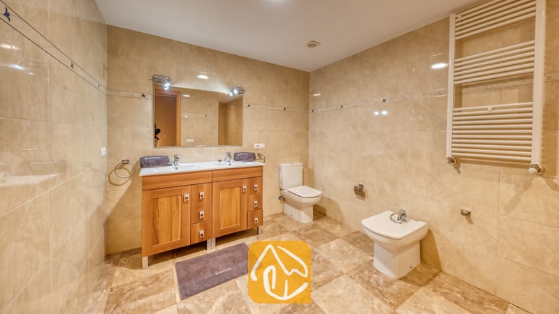 Vakantievilla's Costa Brava Spanje - Villa Grace - Badkamer met beige marmeren tegels op de wanden, een houten wastafelmeubel met dubbele wastafel en een grote wandspiegel, een toilet en een bidet.