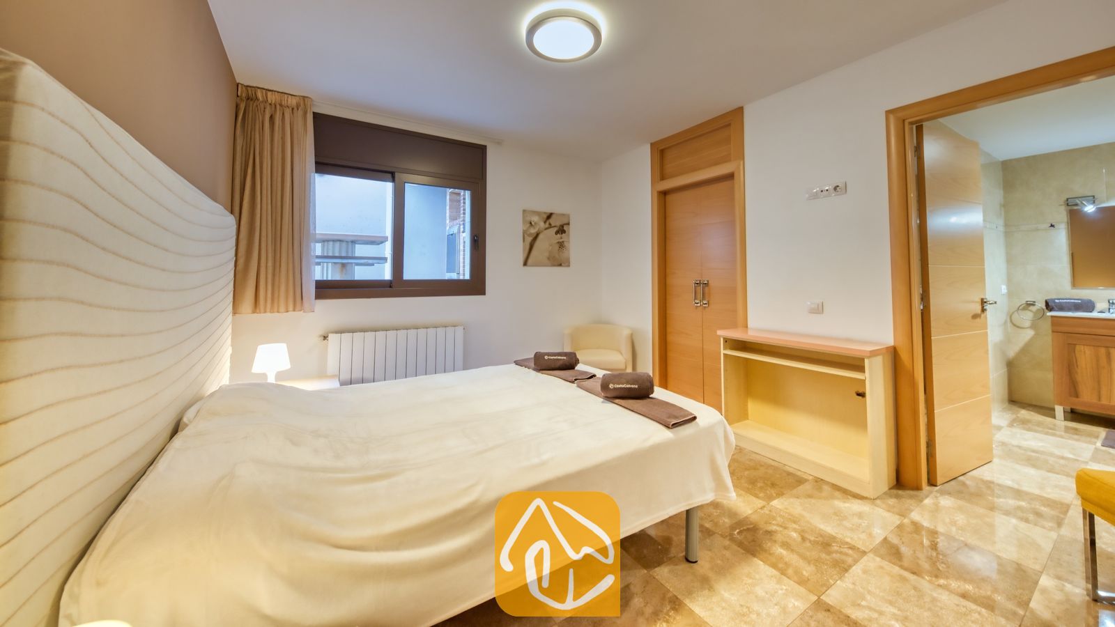 Vakantievilla's Costa Brava Spanje - Villa Grace - Slaapkamer met een wit bed, tegelvloeren, een raam met beige gordijnen en een open doorgang naar een aangesloten badkamer bij Villa Grace.