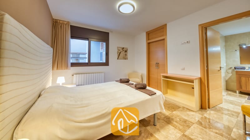 Villas de vacances Costa Brava Espagne - Villa Grace - Chambre avec un lit blanc, un sol carrelé, une fenêtre habillée de rideaux beiges et une porte ouverte menant à une salle de bains attenante à la Villa Grace.