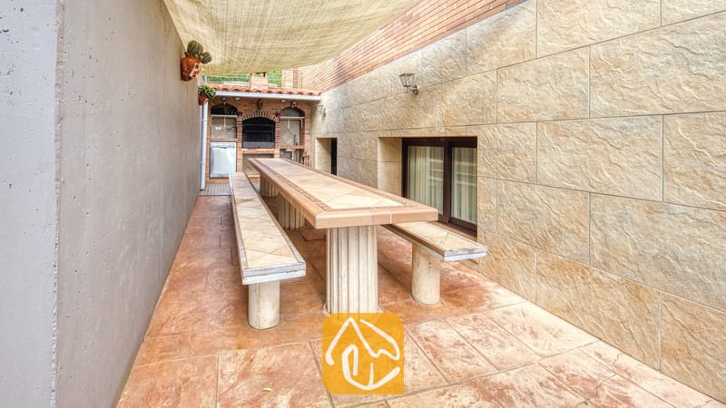 Vakantievilla's Costa Brava Spanje - Villa Grace - Buiten-eethoek bij Villa Grace, voorzien van een lange betegelde tafel met bijpassende banken onder een schaduwdoek, geflankeerd door stenen muren en een stenen barbecue op de achtergrond.