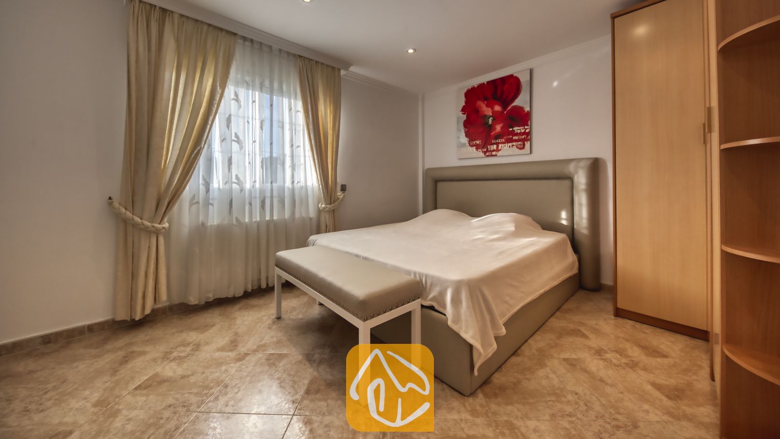 Vakantievilla's Costa Brava Spanje - Villa Lorena - Slaapkamer met een tweepersoonsbed, gestoffeerd bankje, houten kledingkast, tegelvloer en een raam met beige gordijnen.