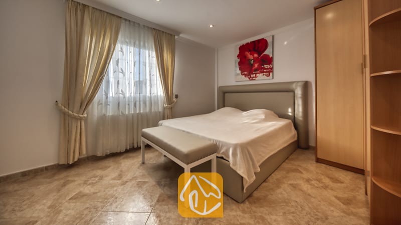 Villas de vacances Costa Brava Espagne - Villa Lorena - Chambre avec un lit double, un banc capitonné, une armoire en bois, un sol carrelé et une fenêtre dotée de rideaux beiges.